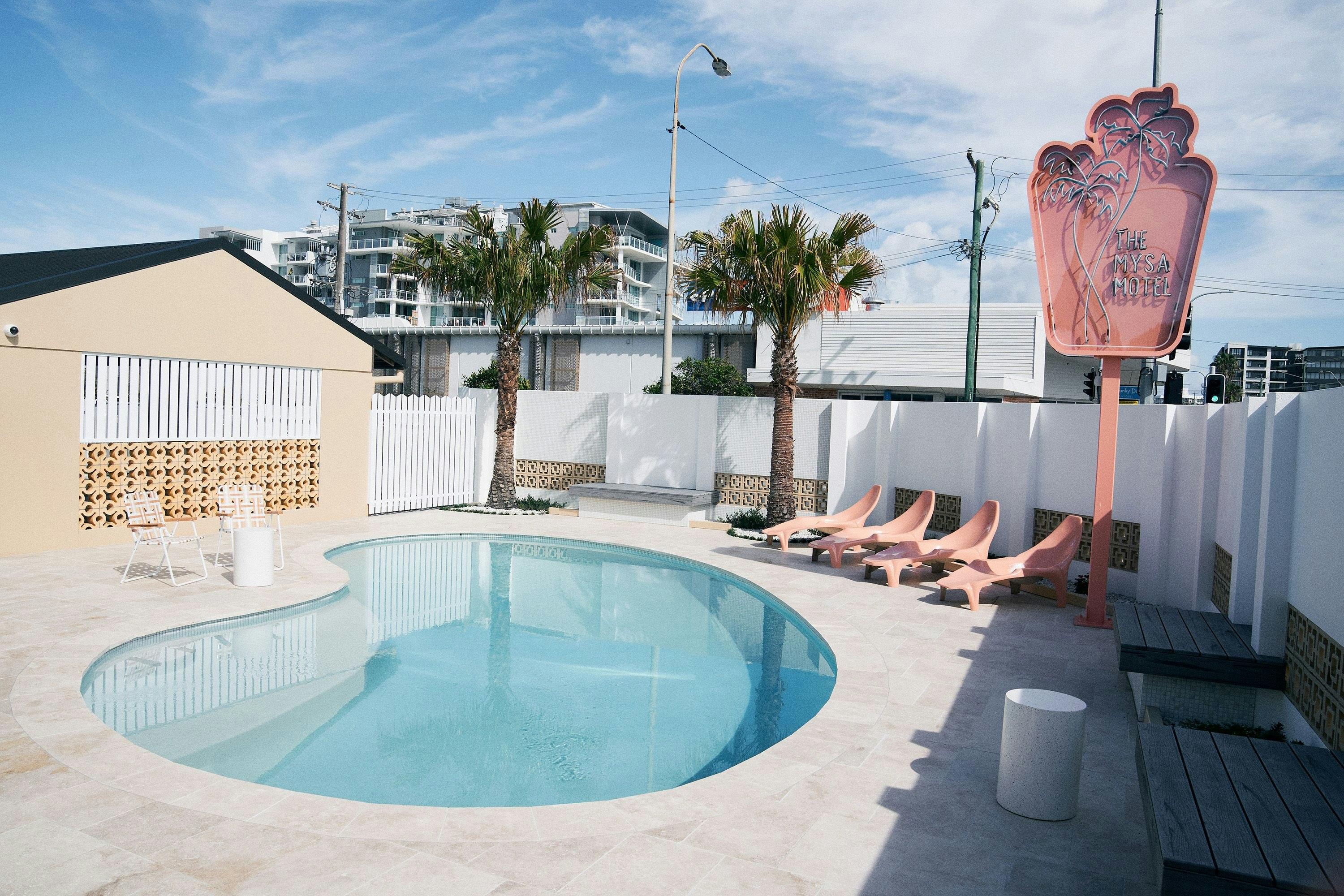the exterior of a retro motel with kidney shaped pool and a neon pink sign and a palm tree